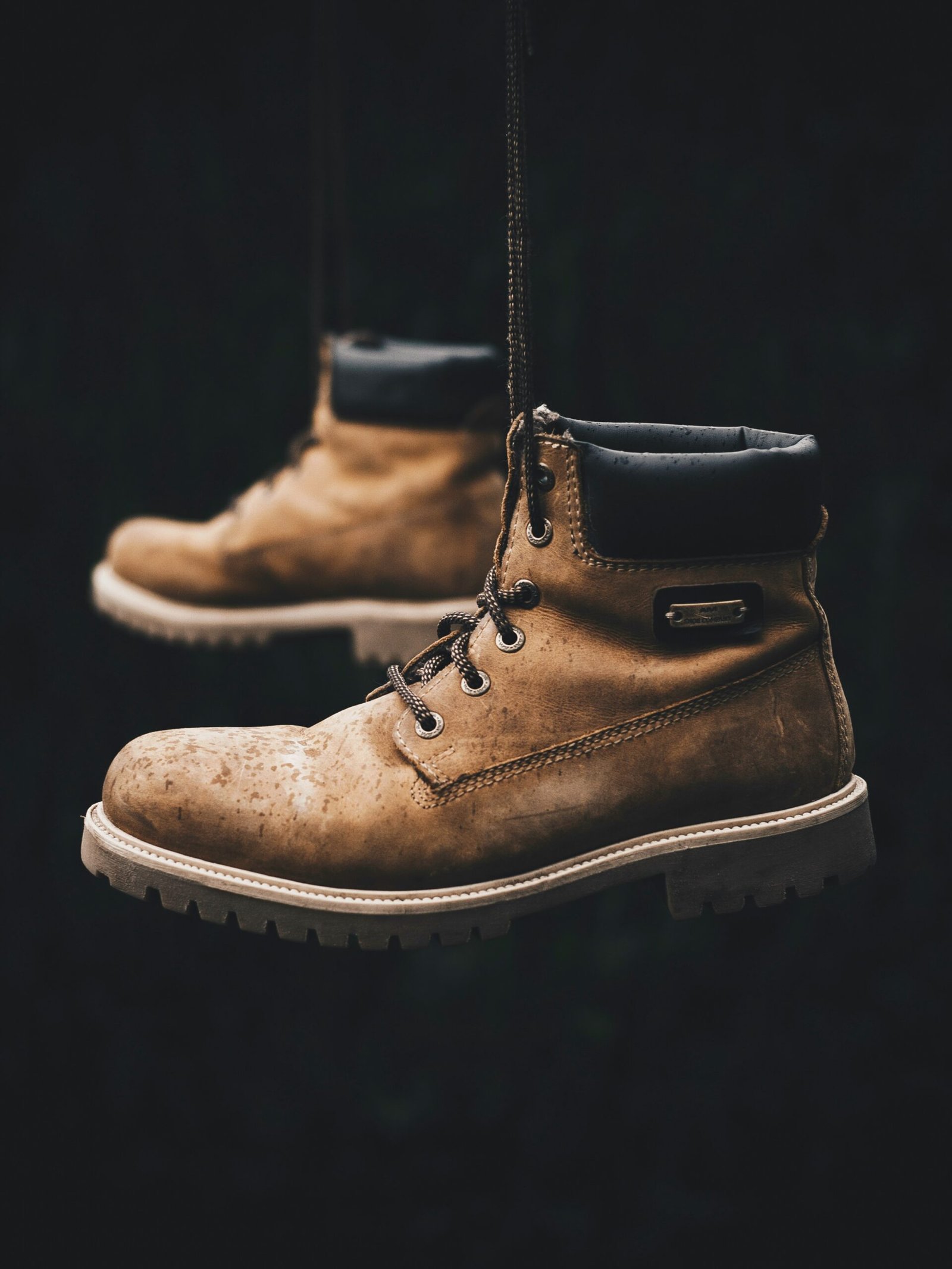 A pair of worn brown leather work boots hanging by their laces against a dark background, with visible scuffs and water stains